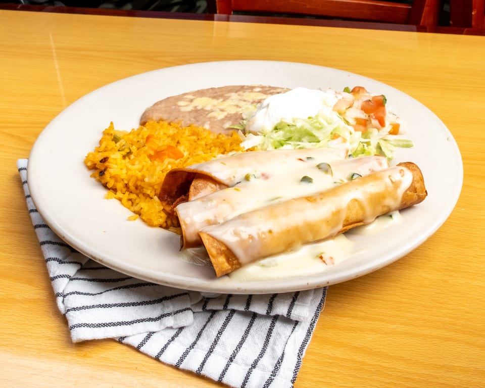 Three taquitos covered in white sauce, served with rice, refried beans, and a side salad.