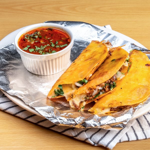 Three cheesy birria tacos with a side of consommé, served on a white plate with foil.