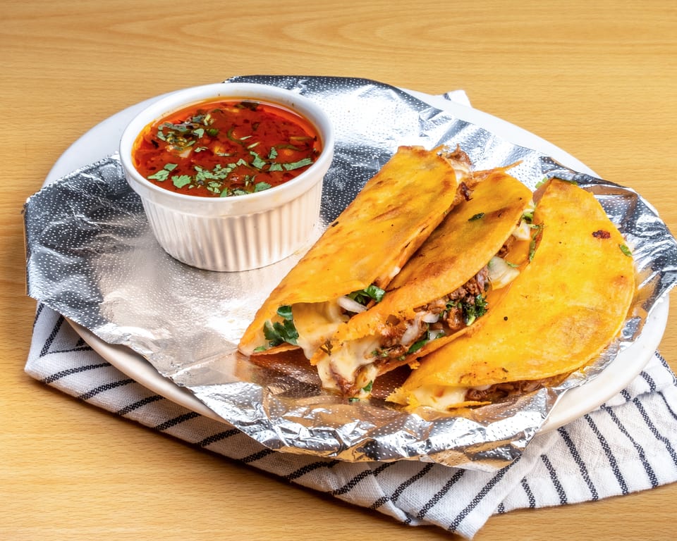 Three cheesy birria tacos with a side of consommé, served on a white plate with foil.