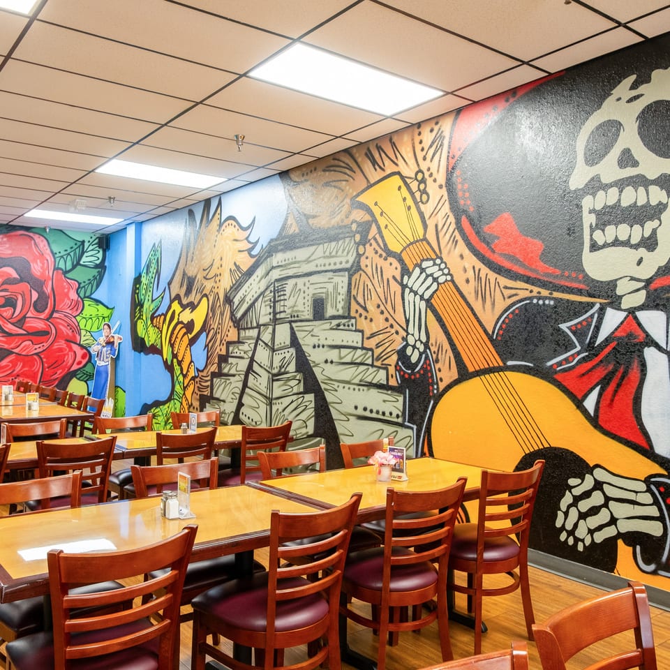 Restaurant dining room with wooden tables and chairs, vibrant murals of a rose, pyramid, and skeleton.