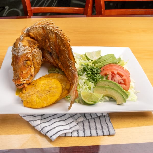 Fried whole fish with plantains, shredded lettuce, avocado, tomato, and lime wedges on a white plate.