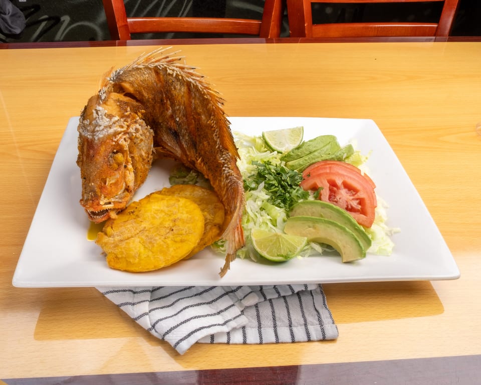 Fried whole fish with plantains, shredded lettuce, avocado, tomato, and lime wedges on a white plate.
