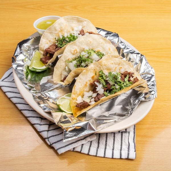 Three street tacos with meat, onions, and cilantro, served with lime wedges and salsa.