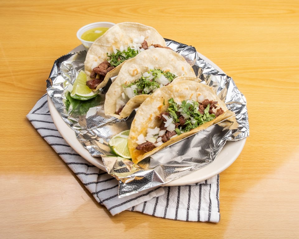 Three street tacos with meat, onions, and cilantro, served with lime wedges and salsa.