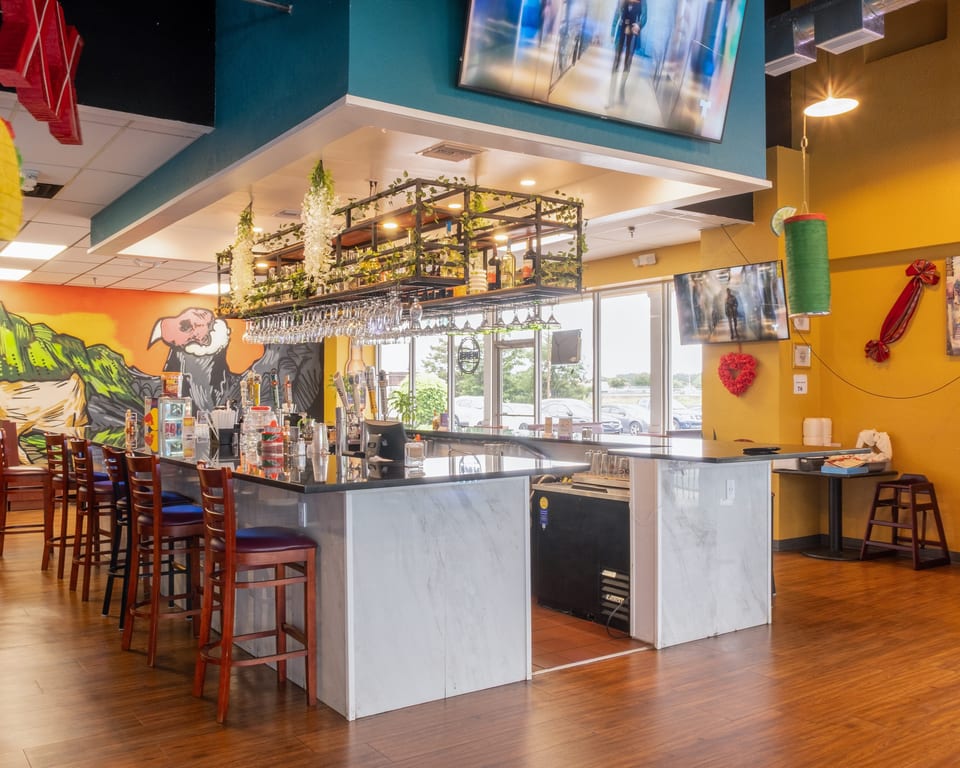 A vibrant restaurant interior features a long bar with stools, a colorful mural, and decorative lighting.