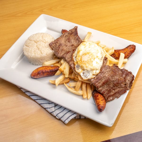 A white rectangular plate holds steak, fried egg, rice, french fries, and plantains on a wooden table.