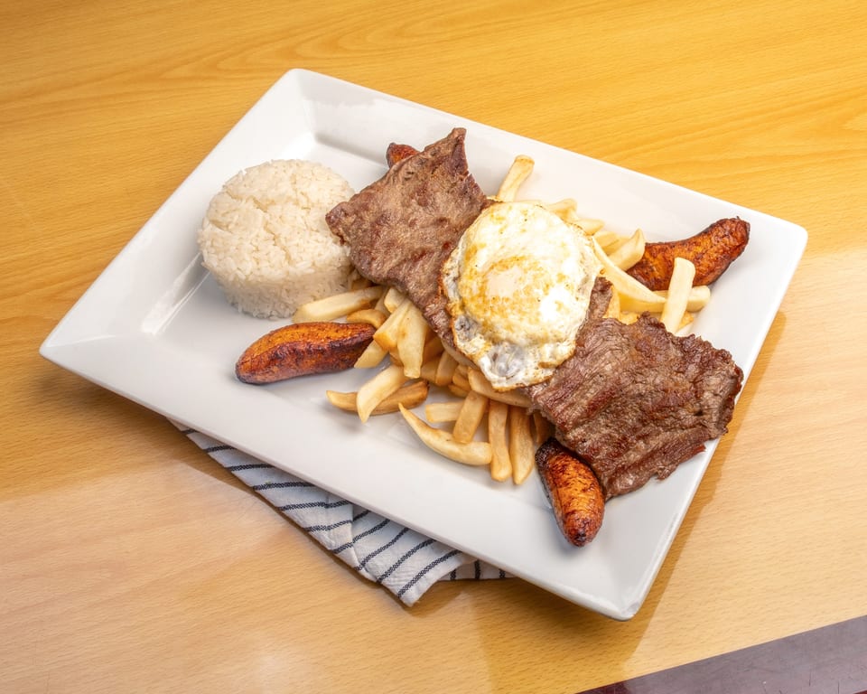 A white rectangular plate holds steak, fried egg, rice, french fries, and plantains on a wooden table.