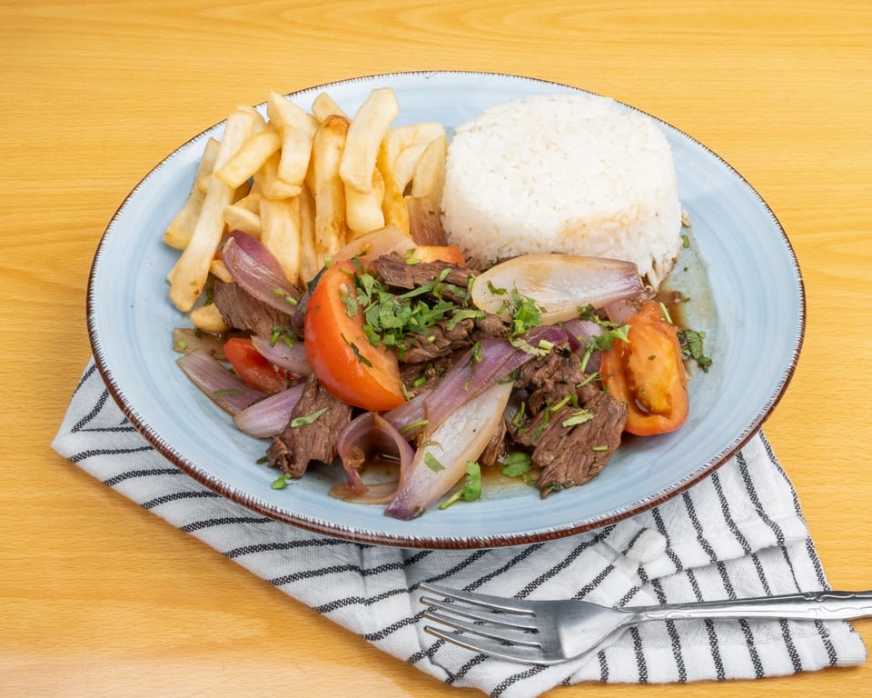 Lomo Saltado with beef, red onions, tomatoes, cilantro, french fries, and white rice on a blue plate.