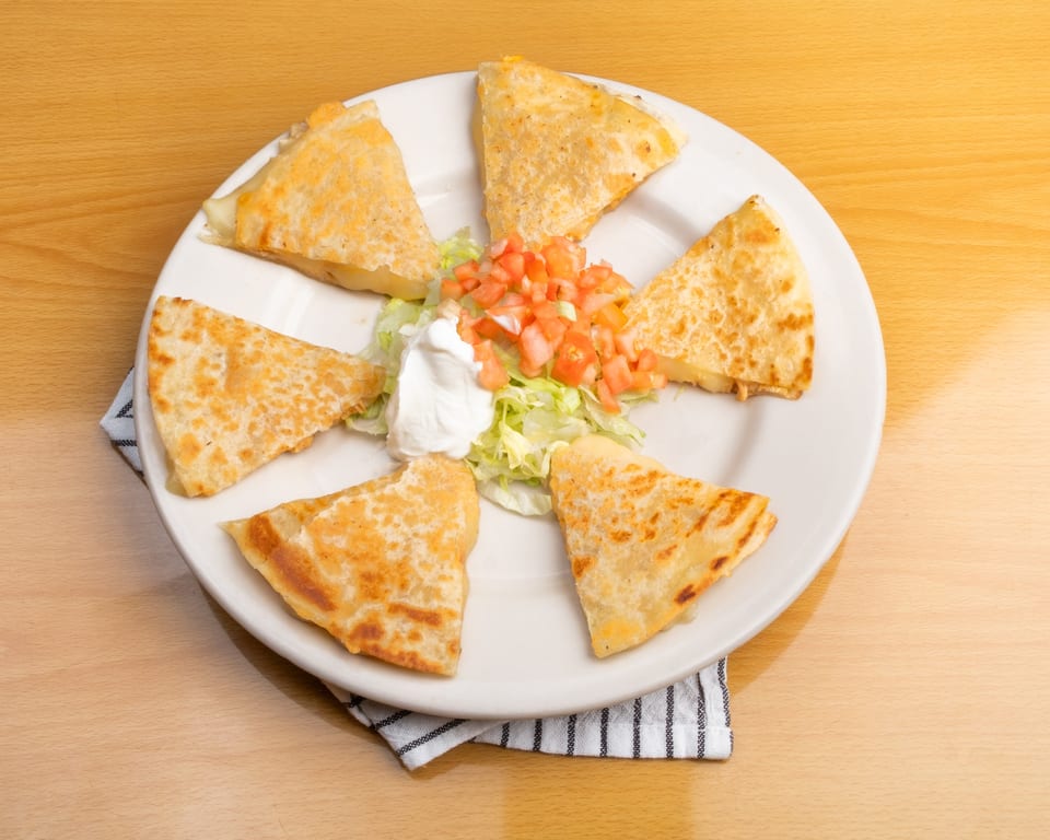 Six quesadilla wedges arranged on a white plate with a side of lettuce, diced tomatoes, and sour cream.