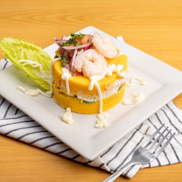 Peruvian causa with yellow mashed potato layers, shrimp, red onion, and cilantro, drizzled with white sauce.