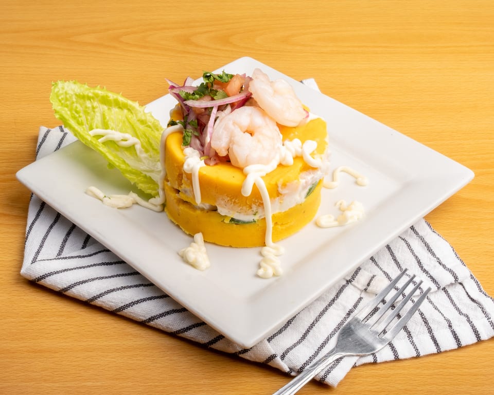 Peruvian causa with yellow mashed potato layers, shrimp, red onion, and cilantro, drizzled with white sauce.